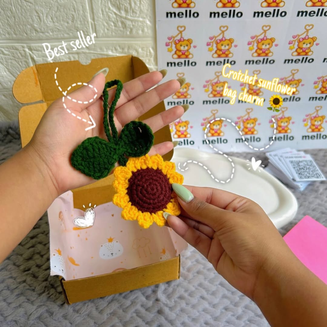 Hand holding a crocheted sunflower bag charm with a branded box in the background.