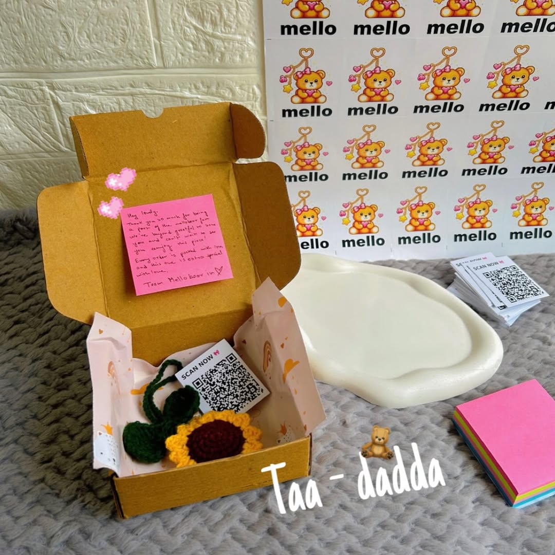 Open cardboard box with a sunflower decoration, pink post-it note, and QR code on a textured surface with 'mello' branding in the background.
