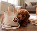 Small brown dog next to a beige handbag with a paw charm on a wooden floor.