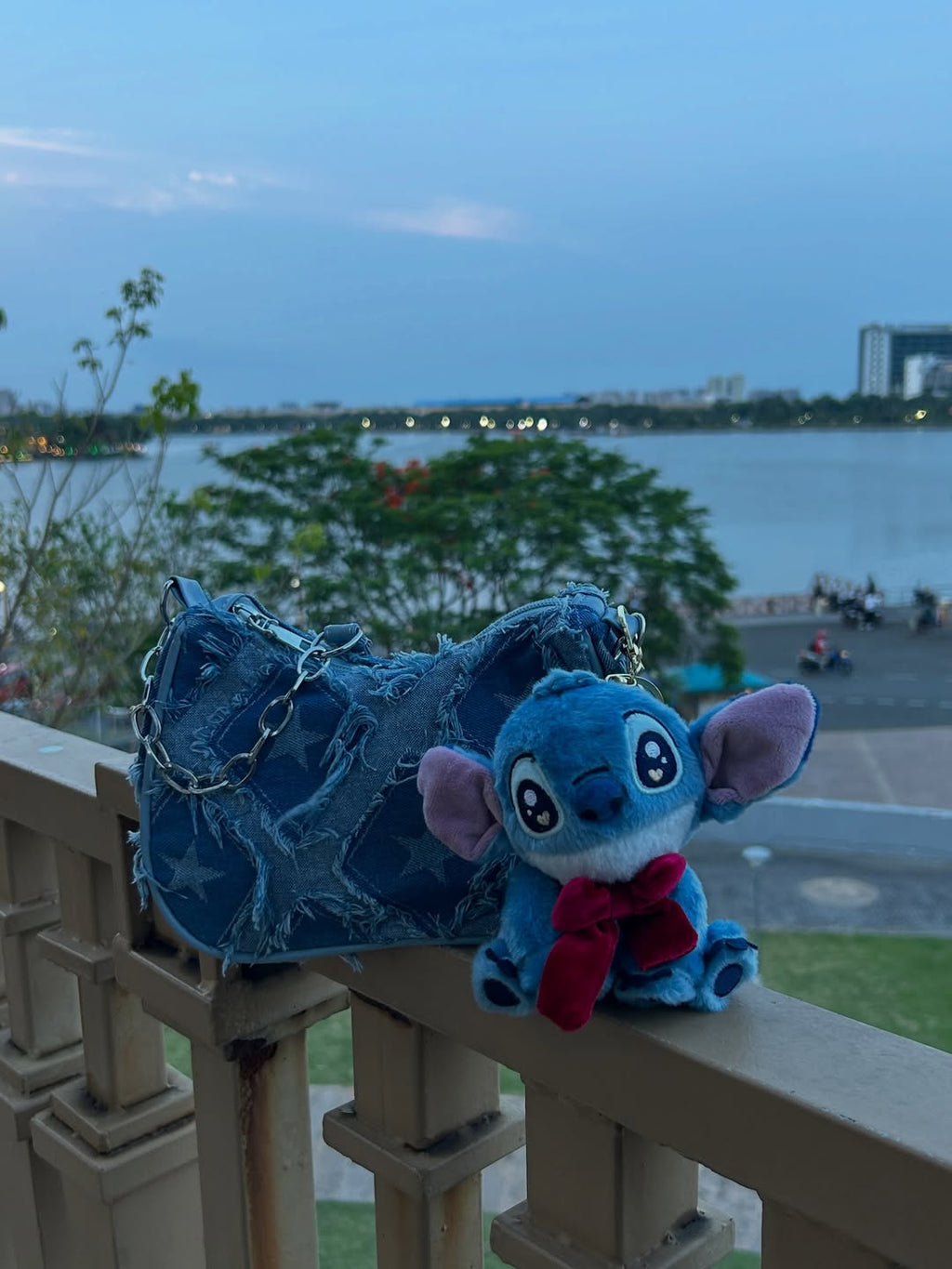 Blue plush toy with a bow tie on a railing with a lake and cityscape in the background