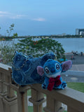 Blue plush toy with a bow tie on a railing with a lake and cityscape in the background