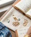 Gold jewelry including a sun-shaped keychain and rings on a textured surface with an open book.